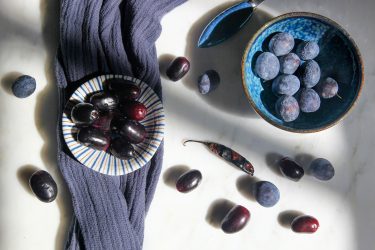 Shiny purple Bangladeshi Jaam fruit in a bowl on the left, and dusky blue Damson plums in a bowl on the right. Ready to make Jaam Bhorta.