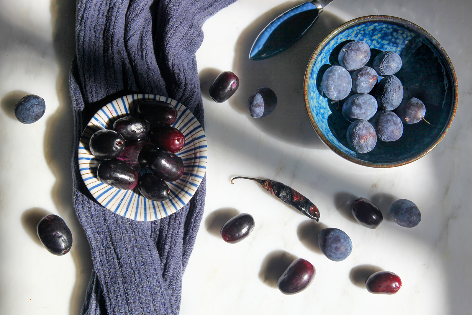 Shiny purple Bangladeshi Jaam fruit in a bowl on the left, and dusky blue Damson plums in a bowl on the right. Ready to make Jaam Bhorta.